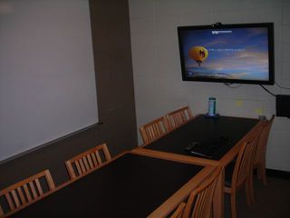 image of study room showing double table with 8 chairs and wall mounted TV/PC on right wall and whiteboard on left wall.